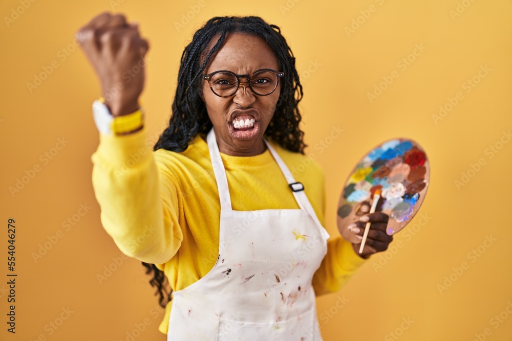 Sticker African woman holding painter palette angry and mad raising fist frustrated and furious while shouting with anger. rage and aggressive concept.