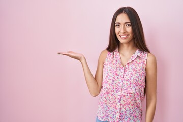 Young hispanic woman with long hair standing over pink background smiling cheerful presenting and pointing with palm of hand looking at the camera.