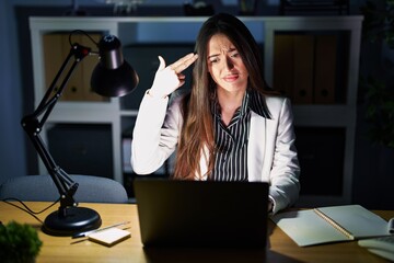 Young brunette woman working at the office at night with laptop shooting and killing oneself pointing hand and fingers to head like gun, suicide gesture.