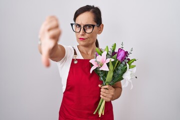 Middle age brunette woman wearing apron working at florist shop holding bouquet looking unhappy and angry showing rejection and negative with thumbs down gesture. bad expression.
