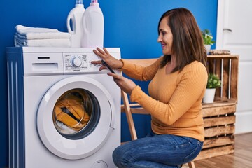 Middle age woman turning on washing machine using smartphone at laundry room