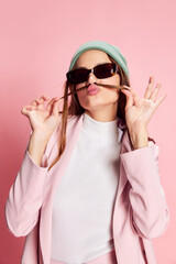 Portrait of young beautiful girl in a suit, hat and sunglasses posing over pink background. Good mood