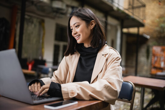 Cheerful asian blogger in trench coat using laptop on terrace of cafe.