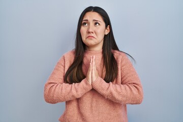 Fototapeta premium Young latin woman standing over blue background begging and praying with hands together with hope expression on face very emotional and worried. begging.