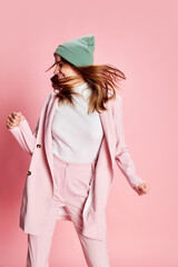 Portrait of young beautiful girl in a suit, hat posing, dancing over pink background. Good mood, positive vibe