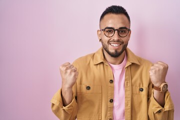 Young hispanic man standing over pink background celebrating surprised and amazed for success with arms raised and open eyes. winner concept.