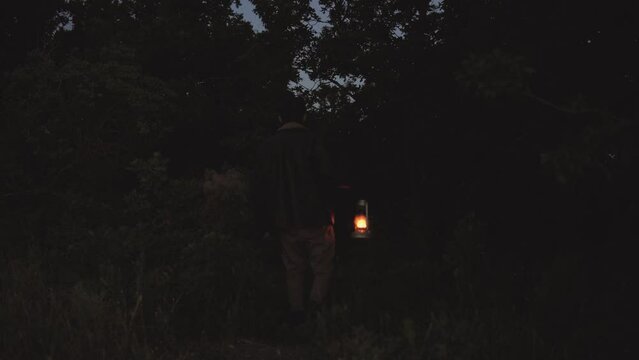 Adult afraid man with beard wear retro clothes travel a night. Seriously guy walking on the dark forest use vintage kerosene lamp try to find exit.