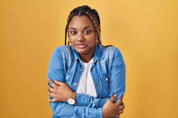 African american woman with braids standing over yellow background shaking and freezing for winter...