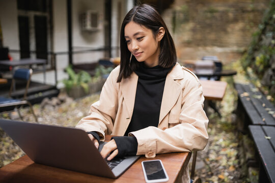 Brunette asian blogger in trench coat using laptop in outdoor cafe.