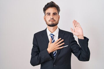 Young hispanic man with tattoos wearing business suit and tie swearing with hand on chest and open...