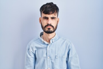 Young hispanic man with beard standing over blue background skeptic and nervous, frowning upset because of problem. negative person.
