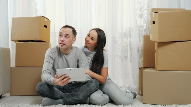 Man and woman sit in new apartment and look at tablet choosing decoration. Excited and amused married couple enjoys moving in new flat and sits among carton boxes