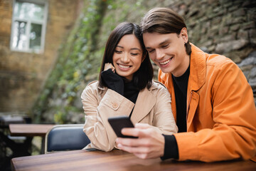 Smiling interracial couple using smartphone in outdoor cafe.