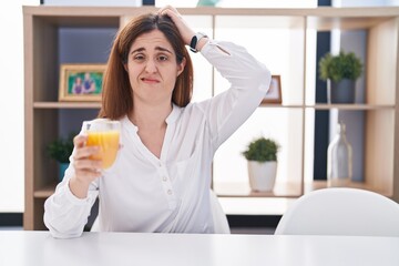 Brunette woman drinking glass of orange juice confuse and wondering about question. uncertain with doubt, thinking with hand on head. pensive concept.