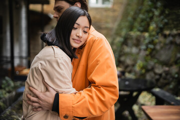 Asian woman hugging stylish boyfriend on terrace of cafe.