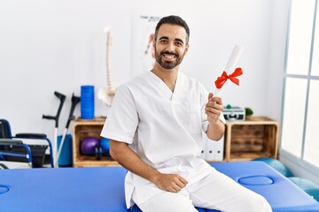 Young hispanic man wearing physiotherapist uniform holding diploma at clinic