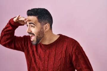 Young hispanic man with beard wearing casual sweater over pink background very happy and smiling looking far away with hand over head. searching concept.