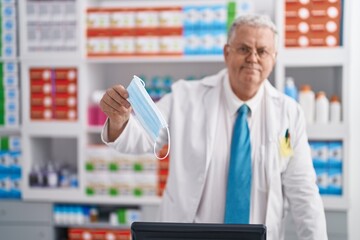 Middle age grey-haired man pharmacist smiling confident holding medical mask at pharmacy