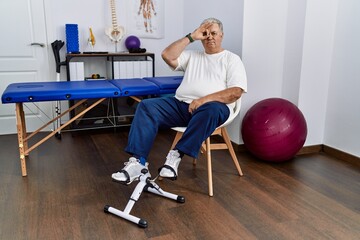 Fototapeta premium Senior caucasian man at physiotherapy clinic using pedal exerciser doing ok gesture with hand smiling, eye looking through fingers with happy face.