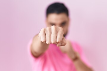 Obraz na płótnie Canvas Hispanic young man standing over pink background punching fist to fight, aggressive and angry attack, threat and violence