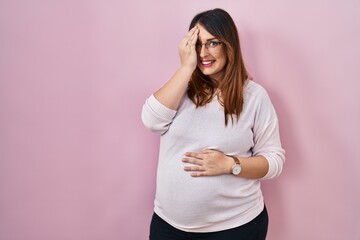 Pregnant woman standing over pink background covering one eye with hand, confident smile on face and surprise emotion.