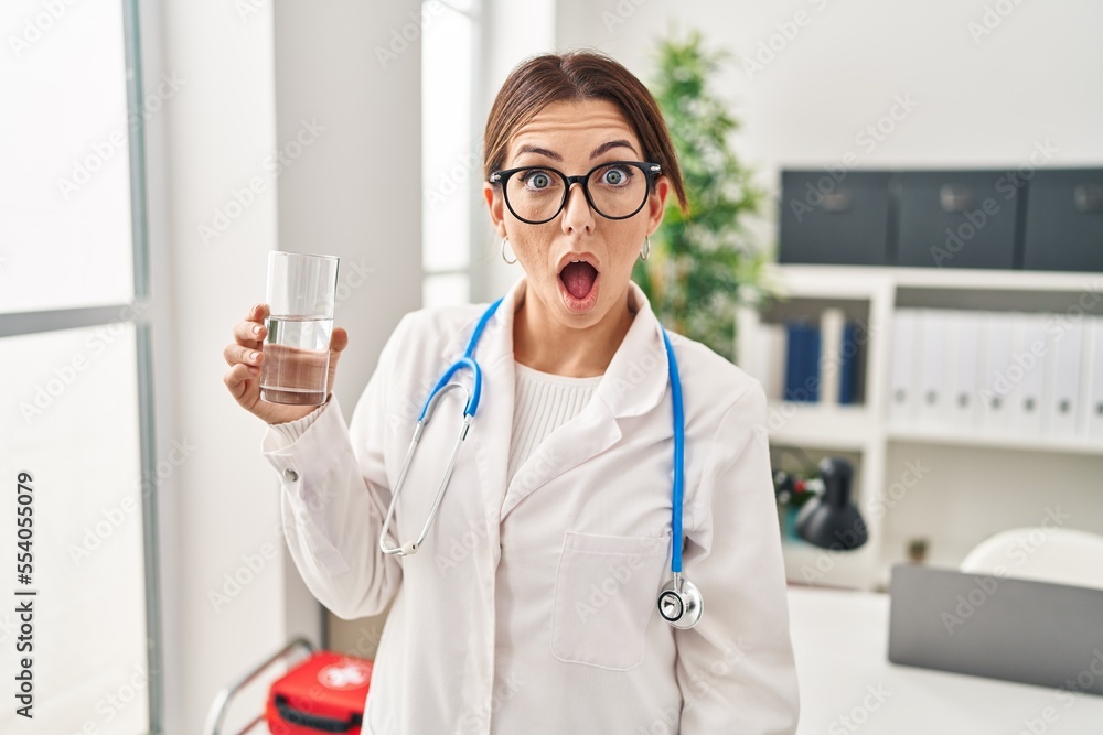 Canvas Prints Young brunette doctor woman holding glass of water scared and amazed with open mouth for surprise, disbelief face