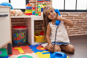 Adorable hispanic girl playing telephone toy sitting on floor at kindergarten