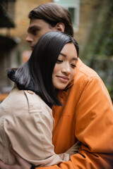 Young asian woman in trench coat hugging blurred boyfriend on terrace of cafe.