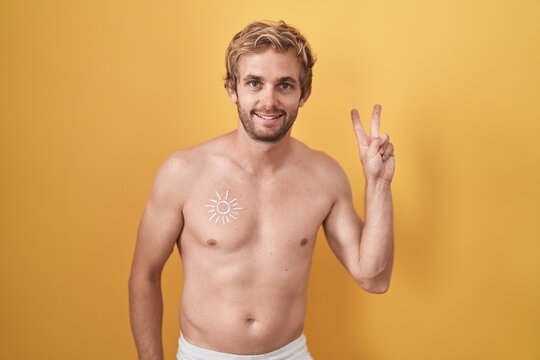 Caucasian man standing shirtless wearing sun screen smiling with happy face winking at the camera doing victory sign with fingers. number two.