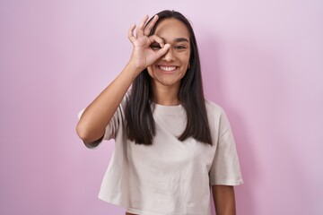 Obraz na płótnie Canvas Young hispanic woman standing over pink background doing ok gesture with hand smiling, eye looking through fingers with happy face.