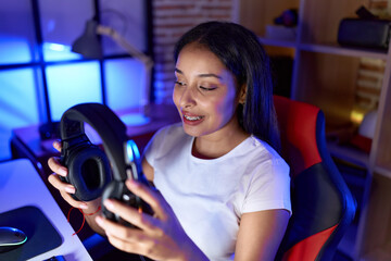 Fototapeta premium Young arab woman streamer smiling confident holding headphones at gaming room