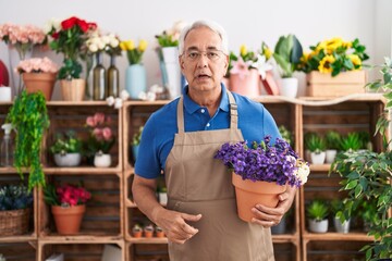 Middle age man with grey hair working at florist shop holding plant scared and amazed with open mouth for surprise, disbelief face