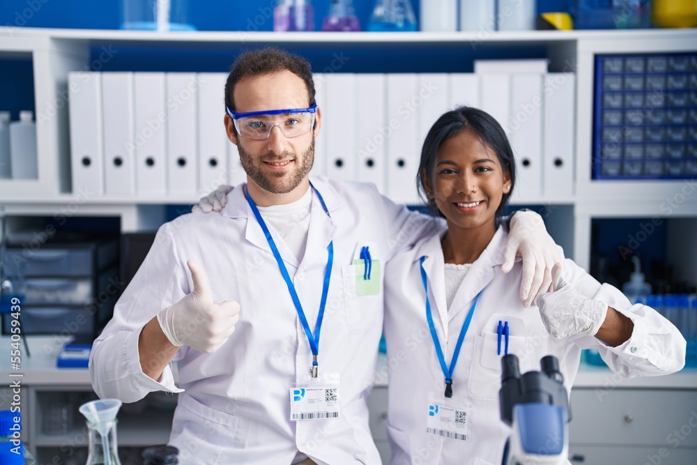 Wall mural Interracial couple working at scientist laboratory smiling happy and positive, thumb up doing excellent and approval sign