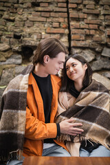 Fototapeta na wymiar Smiling interracial couple in blanket looking at each other while sitting in blanket in outdoor cafe.