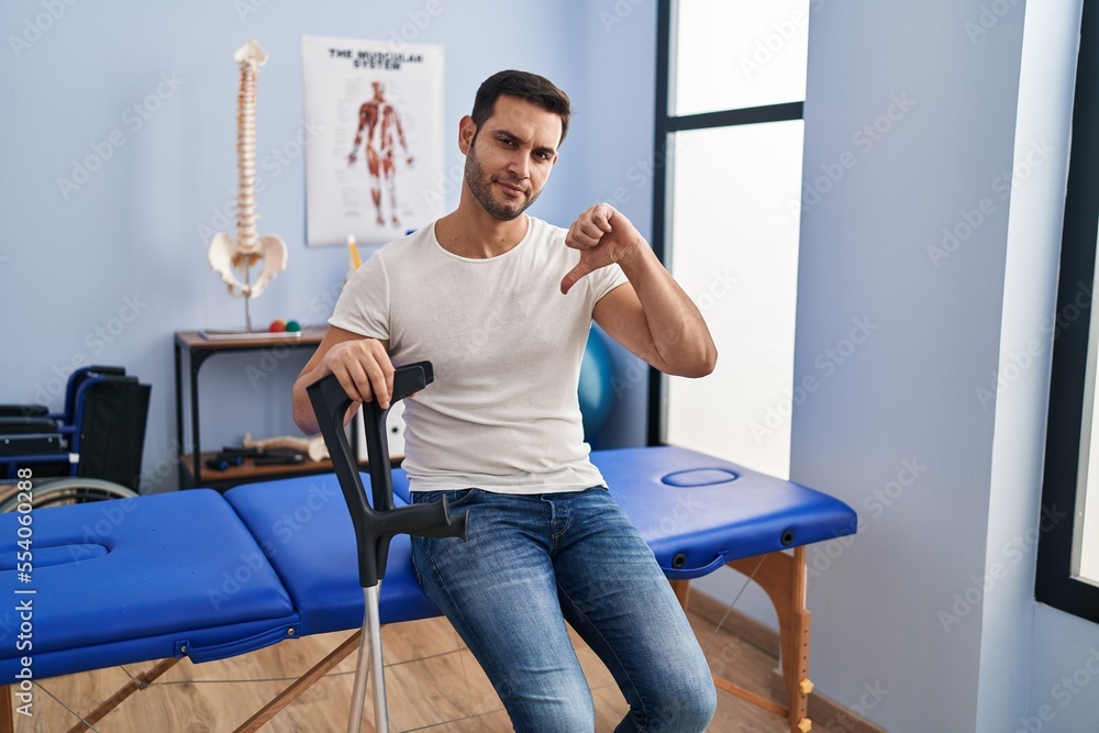 Sticker Young hispanic man with beard wearing crutches at rehabilitation clinic with angry face, negative sign showing dislike with thumbs down, rejection concept