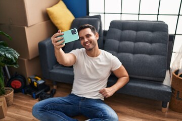Young hispanic man make selfie by smartphone sitting on floor at new home