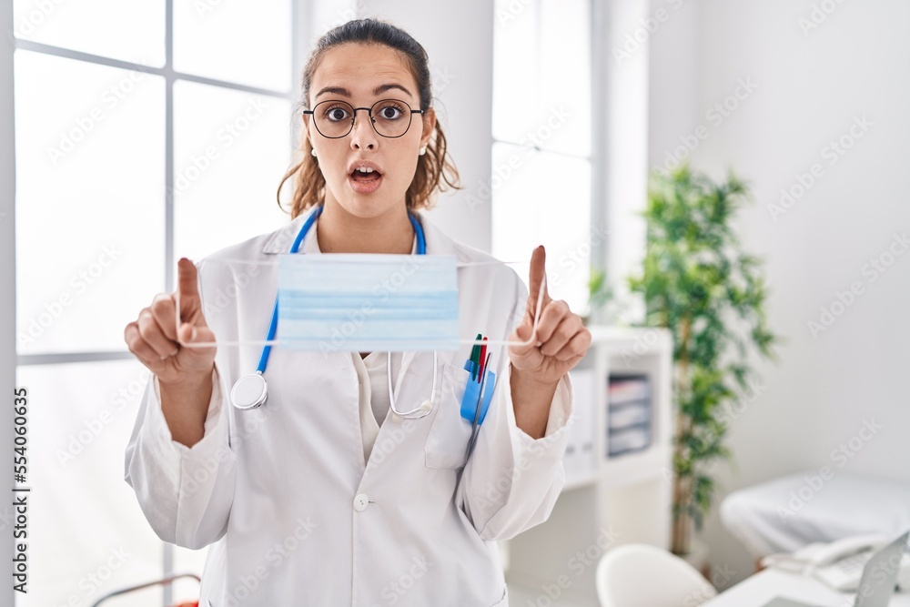 Sticker Young hispanic doctor woman holding safety mask in shock face, looking skeptical and sarcastic, surprised with open mouth