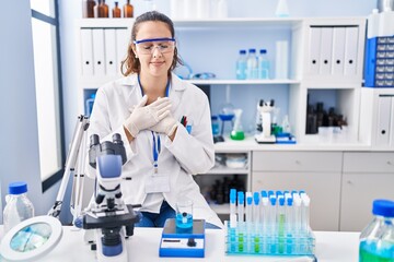 Fototapeta premium Young hispanic woman working at scientist laboratory smiling with hands on chest with closed eyes and grateful gesture on face. health concept.