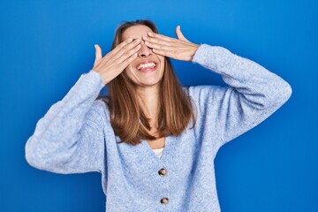 Young woman standing over blue background covering eyes with hands smiling cheerful and funny....