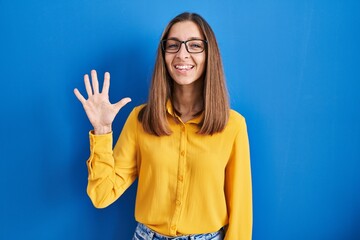 Young woman wearing glasses standing over blue background showing and pointing up with fingers number five while smiling confident and happy.