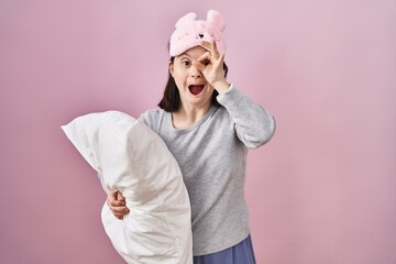 Woman with down syndrome wearing sleeping mask hugging pillow doing ok gesture shocked with surprised face, eye looking through fingers. unbelieving expression.