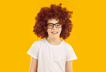 Portrait of happy child model in fashion studio. Cheerful little boy in funny big brown curly wig, white T shirt and glasses standing isolated on orange color background