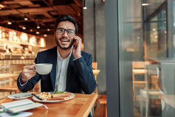 Handsome indian businessman in glasses is talking phone with client during lunch time in cozy cafe