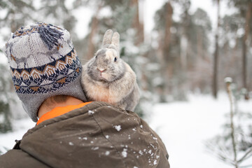 Close portrait of a man with a rabbit. The guy gently hugs a gray fluffy bunny in winter snowy forest. Travel with pet. 