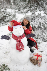 Asian girl building snowman in snowy park. Woman embracing snowman wearing hat and scarf. Active outdoors leisure in winter park. 
