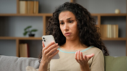 Hispanic sad displeased woman has problem with online mobile app on cell phone unhappy with blocked account in social networks looking at cellphone screen scam unsuccessful bad news sitting at home