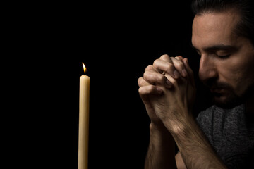 A man folded his hands in prayer next to a burning candle on a black background. prayer to God for...