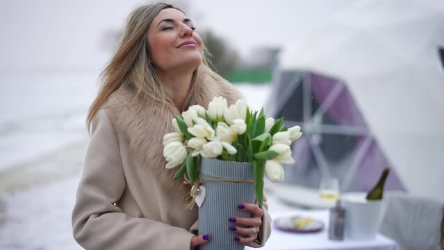 Portrait of confident gorgeous woman smelling bouquet of white flowers smiling standing on snowy day outdoors. Live camera zoom in to satisfied Caucasian lady with tulips on Valentine's Day