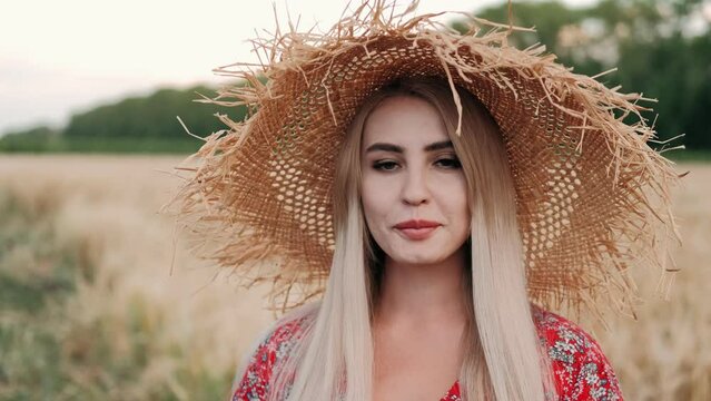 Portrait of a young beautiful girl in a wide straw hat at sunset looks at the camera and smiles. Young Woman posing on a sunny warmly Day. Happiness concept. High quality 4k footage