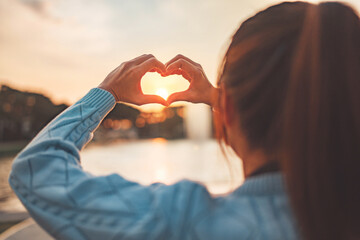 Hand of woman showing heart love symbol shape with sunrise, Happy travel and freedom in nature concept.
